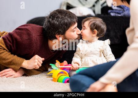 Loving bearded father kissing cute little daughter playing with toys near crop mother sitting on carpet at home Stock Photo