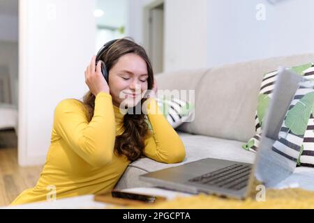 Delighted young female freelancer in yellow turtleneck listening to music in wireless headphones with closed eyes while working on laptop remotely fro Stock Photo