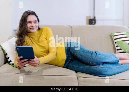 Cheerful young female in casual clothes smiling and browsing tablet while lying on sofa in living room looking away Stock Photo