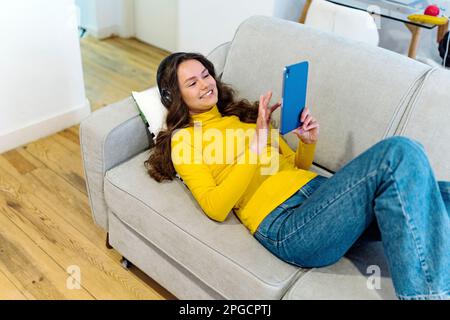 From above happy young female in yellow turtleneck smiling and browsing on tablet while listening to music in headphones during rest on sofa Stock Photo