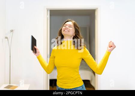 Delighted young female in yellow turtleneck smiling with eyes closed and holding tablet while standing celebrating victory with raised arm at home Stock Photo