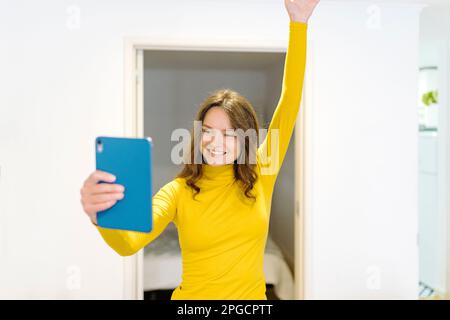 Delighted young female in yellow turtleneck smiling and having video call on tablet while standing with raised arm at home Stock Photo