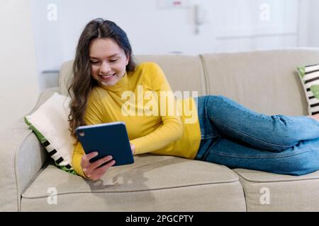 Cheerful young female in casual clothes smiling and browsing tablet while lying on sofa in living room Stock Photo