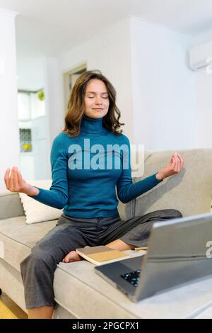 Relaxed young female freelancer in casual clothes sitting on sofa with laptop and meditating with closed eyes while having break during remote work Stock Photo