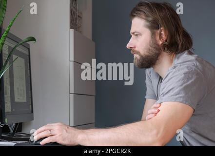 Man working on desktop computer at home. Handsome businessman or freelancer in casual clothes working remotely from home. Pensive young Caucasian man Stock Photo
