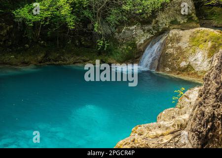 Small waterfall flowing into blue lake surrounded by rocks and green trees growing in forest on sunny day Stock Photo