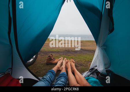 Anonymous traveler couple lying in tent with vehicle parked outside and relaxing during daytime against far away ocean blue sky mountains in Ireland Stock Photo
