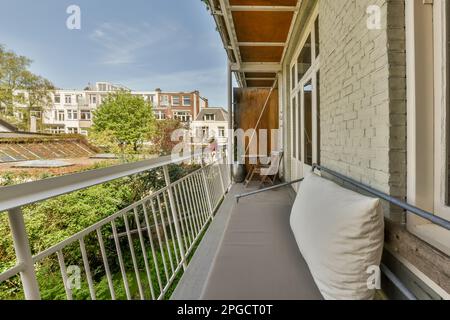 Narrow long balcony with metal railings wooden table and chairs near sofa against brick white wall with windows on sunny day Stock Photo