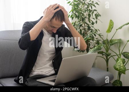 Confused man sitting on couch with computer on lap in living room, upset frustrated male reading bad news on message, having problem with device or Stock Photo
