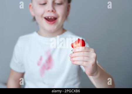 A child's hand is holding a slice of strawberry. A dirty pink stain on the front of a white t-shirt. daily life stain concept Stock Photo