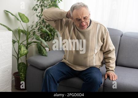 Senior man having headache and touching his head while suffering from a migraine in the living room, Mature man presses a hand to head suffers from Stock Photo