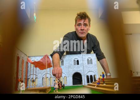 22 March 2023, Saxony-Anhalt, Magdeburg: Moritz Sostmann, director of the play 'The Dragon' stands behind a model of the cloister of the former monastery in the art museum Kloster Unser Lieben Frauen in which figures, a model stage and a model tribune can be seen. The model illustrates the stage and tribune of the upcoming 'Hofspektakel' by the Puppentheater Magdeburg, which will be performed this year in cooperation with the Kunstmuseum Kloster Unser Lieben Frauen. The stage from the puppet theater will be located in the courtyard of the former monastery this season. The fairy tale comedy 'Th Stock Photo