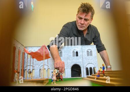 22 March 2023, Saxony-Anhalt, Magdeburg: Moritz Sostmann, director of the play 'The Dragon' stands behind a model of the cloister of the former monastery in which figures, a model stage and a model tribune can be seen in the Kunstmuseum Kloster Unser Lieben Frauen. The model illustrates the stage and tribune of the upcoming 'Hofspektakel' by the Magdeburg Puppet Theater, which will be performed this year in cooperation with the Unser Lieben Frauen Monastery Art Museum. The stage from the puppet theater will be located in the courtyard of the former monastery this season. The fairy tale comedy Stock Photo