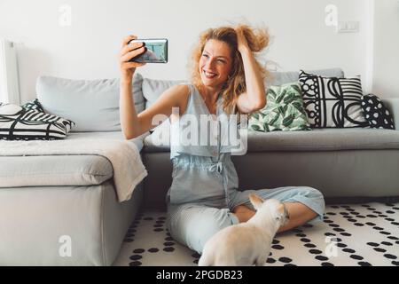 Smiling curly woman playing with her hair while taking selfies with her phone, sitting on the carpet by the couch Stock Photo