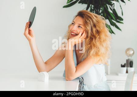 Smiling blonde woman with curly hair looking at herself in a small round mirror she is holding up Stock Photo