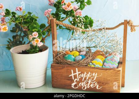 Wooden basket with Easter eggs and sweets on the table. An ordinary kitchen room with flowers in a pot. Easter concept. Stock Photo
