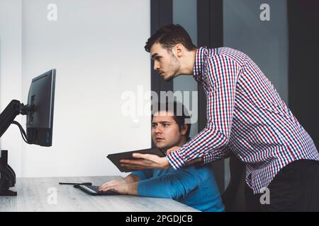Two young IT programmers coding, working on a joint project in the office, one holding a digital tablet while other works on desktop computer  Stock Photo