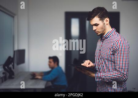 Side view of young programmer standing in the office looking down at the digital tablet he is holding in his hands  Stock Photo
