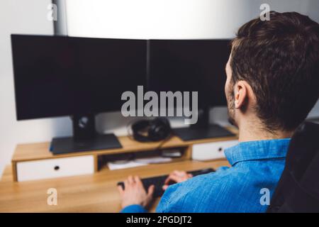 Over the shoulder view unrecognizable man in blue shirt working on two black screens  Stock Photo