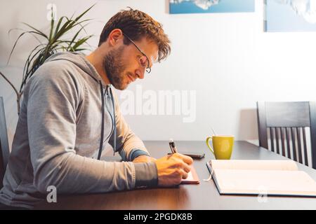 Side view of man with glasses wearing a tracksuit, sitting by the table making notes studying at home Stock Photo