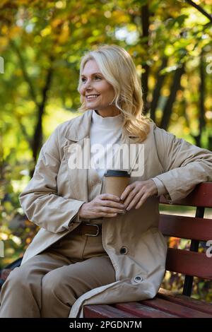 Carefree blonde woman holding paper cup while sitting on bench in spring park,stock image Stock Photo