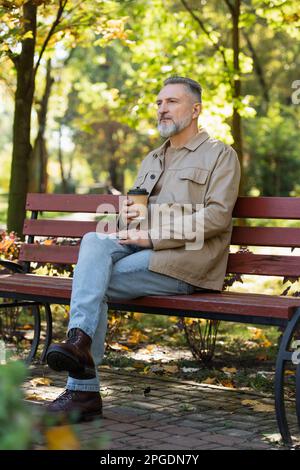 Mature man holding coffee to go while sitting on bench in park,stock image Stock Photo