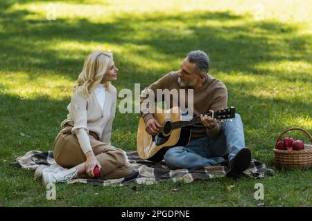 middle aged man with grey beard playing acoustic guitar near charming wife during picnic in park,stock image Stock Photo