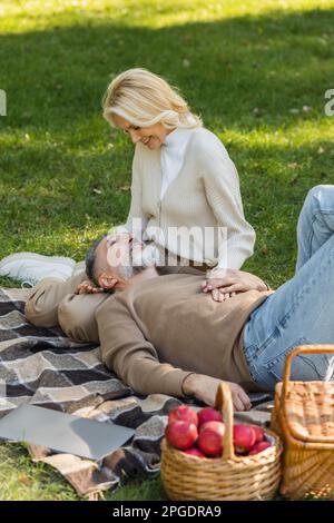 pleased middle aged man lying on laps of blonde and cheerful wife during picnic in park,stock image Stock Photo