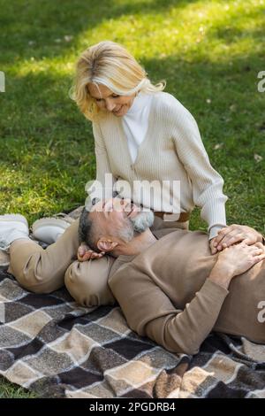 pleased middle aged man lying on laps of blonde and happy wife during picnic in park,stock image Stock Photo