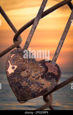 Close-up of a rusty heart shaped padlock on a metal fence by the ocean at sunset, USA Stock Photo