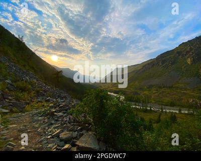 Trail along the mountain near the valley under the sunlit mountain at sunset with the Chuya river under the stone cliff under the clouds in the shade Stock Photo