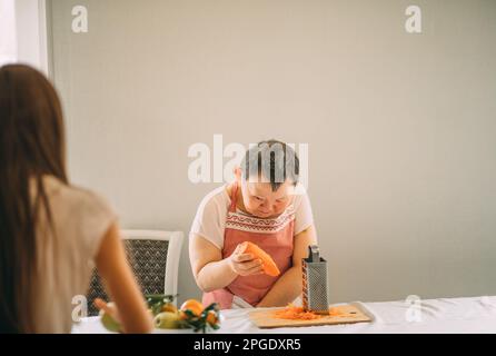 Lifestyle, education. An elderly woman with Down syndrome rubs carrots on a grater with an assistant Stock Photo