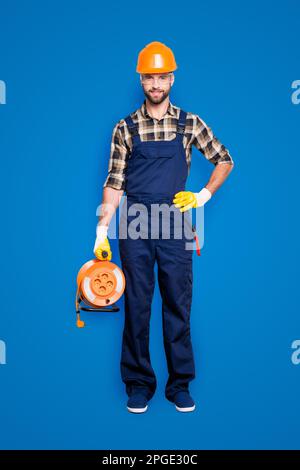 Full size fullbody portrait of cheerful joyful electrician with bristle in overall, shirt, holding equipment with rolled cable in hand and arm on Stock Photo
