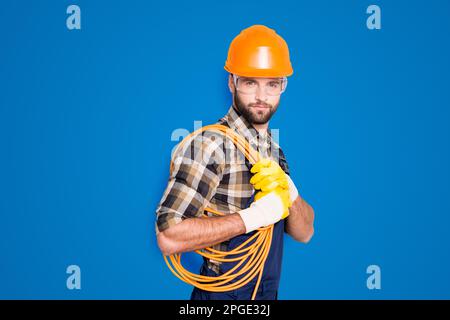 Portrait of trendy virile electrician in hardhat, overall, shirt is ready for work, having, holding rolled cable on shoulder, looking at camera Stock Photo