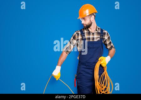 Half face portrait of busy stylish electrician with stubble in overall, shirt, hardhat installing, laying a cable, having rolled wire in hand Stock Photo