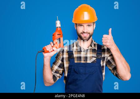 Portrait of joyful cheerful repairer in protective hard hat, overalls, shirt with bristle showing thumb up having equipment in arm isolated on grey Stock Photo