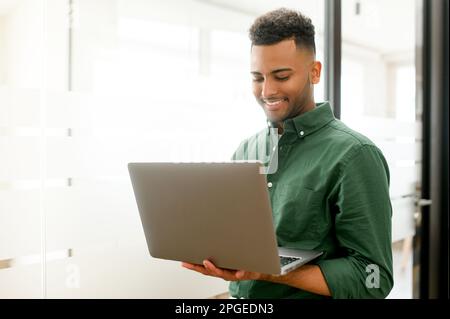 Smiling freelancer guy wearing casual shirt using laptop in contemporary office space, concentrated man typing, messaging, indian programmer develops software, looks at computer screen Stock Photo