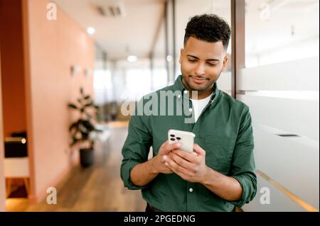 Handsome hispanic guy is using smartphone for messaging, texting, young indian man in smart casual green shirt using smartphone standing in office, typing message, chatting in the networks, scrolling Stock Photo