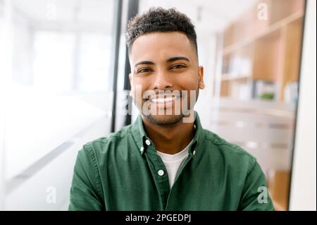 Close-up portrait of a happy indian young man with friendly wide toothy smile, latin guy wearing casual ggreen shirt looks into camera, employee profile photo Stock Photo