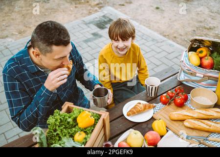 Father dad and school kid boy child eating croissant picnic forest camping site with vegetables, juice, coffee, and croissants. Wooden crate with Stock Photo