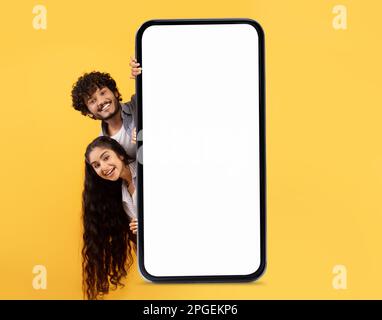Nice Offer. Happy Indian Couple Standing Behind Blank Smartphone With White Screen Stock Photo