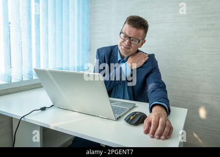 man with stiff shoulder in front laptop. health problems from sedentary work. a man in a suit is holding on to a sore neck Stock Photo