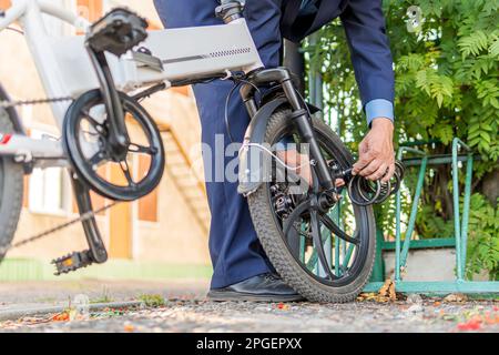 image of the hand locking the bicycle lock. An employee in a suit or a businessman is strapping on his electric bike near work. Stock Photo