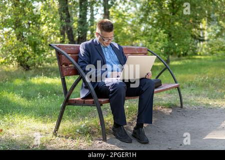 Handsome businessman in blue suit sitting on the bench in summer park working remotely on laptop. businessman is resting with a laptop in the park in Stock Photo