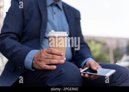 business, education, technology, communication and people concept - creative man with smartphone drinking coffee from paper cup and sitting on city st Stock Photo