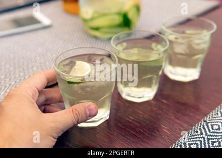 A glass with lemonade, lemon wedges, ice cubes. Summer cold refreshing drink in the hand of a man on the table in a cafe. glasses of cool refreshing l Stock Photo
