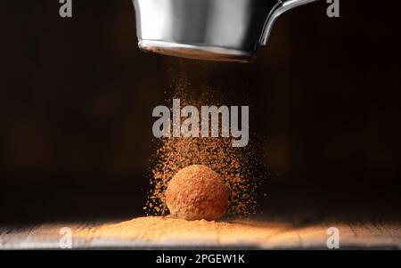 A ball of chocolate truffle is sprinkled with cocoa from a strainer on a dark background close-up. Chocolate Fiery. Conceptual photo Stock Photo