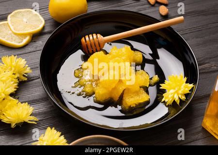 Crystallized honey, dipper in a black plate on a dark wooden background. Composition of honey, lemon, yellow flowers and petals on a black background. Stock Photo