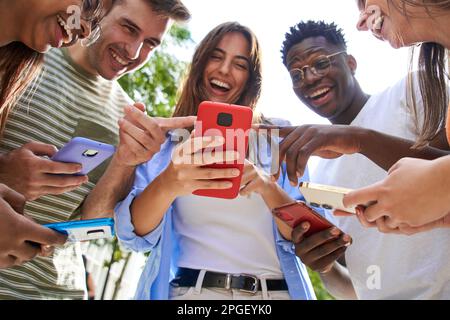 Low angle view of group smiling multiracial young people with cell watching funny reels on mobile. Stock Photo