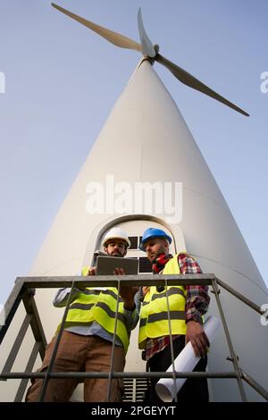 Vertical two helmeted engineers using and looking tablet. Professional technical at wind power plant Stock Photo